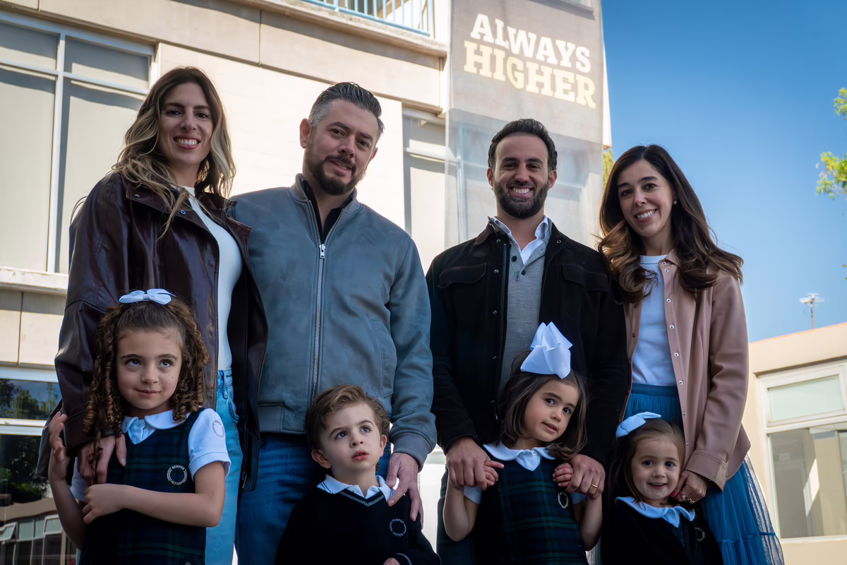 Dos parejas adultas sonrientes con cuatro niños en uniformes escolares frente a un edificio con el letrero 'ALWAYS HIGHER'.